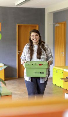 Eine Frau lächelt, während sie eine grüne Kiste in einem modernen Büro mit hellen Farben trägt. An den Wänden sind „#TEAM MEMO” und „CONTAINER PARKPLATZ” zu sehen.