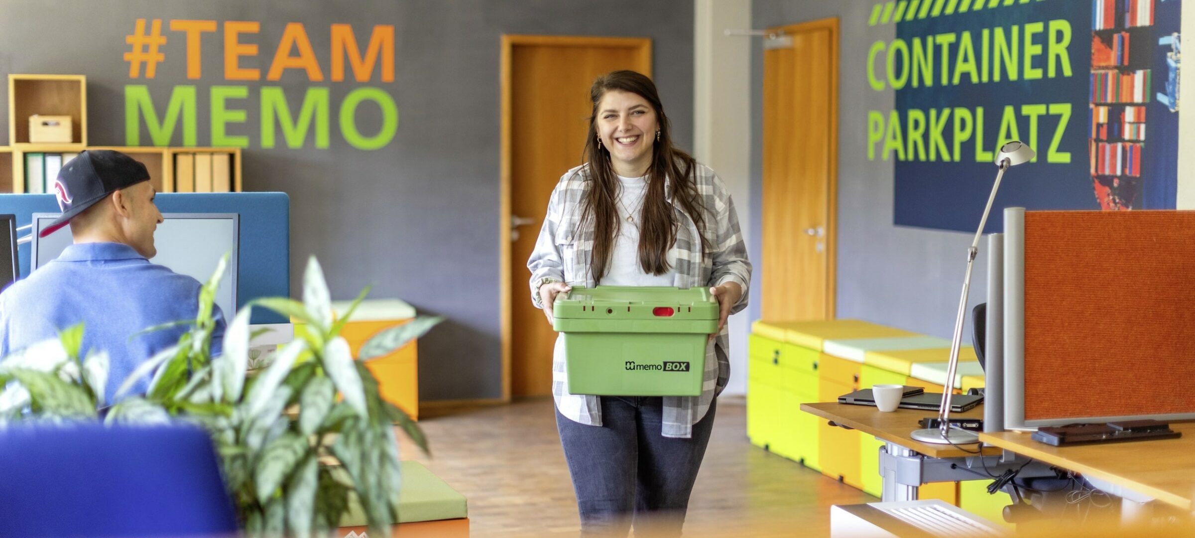 Eine Frau lächelt, während sie eine grüne Kiste in einem modernen Büro mit hellen Farben trägt. An den Wänden sind „#TEAM MEMO” und „CONTAINER PARKPLATZ” zu sehen.
