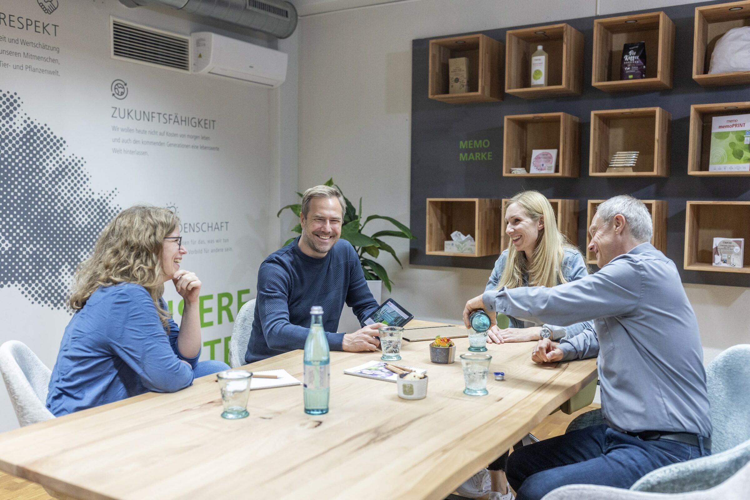 Vier Personen vom Team der nachhaltigen memo AG sitzen um einen Holztisch herum und führen ein konstruktives Gespräch. Im Hintergrund sind Regale mit verschiedenen Gegenständen zu sehen, die eine kreative Atmosphäre schaffen.