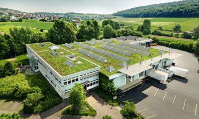 Aerial view of memo AG's headquarters with a green roof and solar panels, surrounded by lush greenery and fields. The scenery is peaceful, with hills in the distance and a small village.