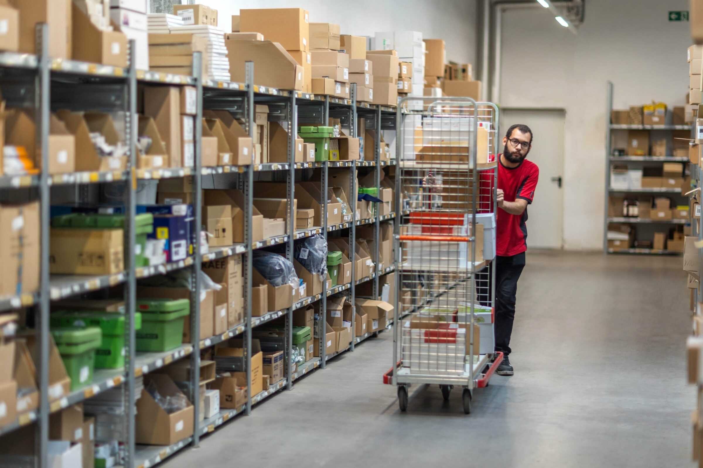 Ein Mann in einem roten Hemd schiebt einen mit Kartons beladenen Wagen durch einen Lagergang des nachhaltigen Versandhandels memo. An den Seiten stehen Regale voller Kartons und Gegenstände, die eine geschäftige Arbeitsumgebung vermitteln.