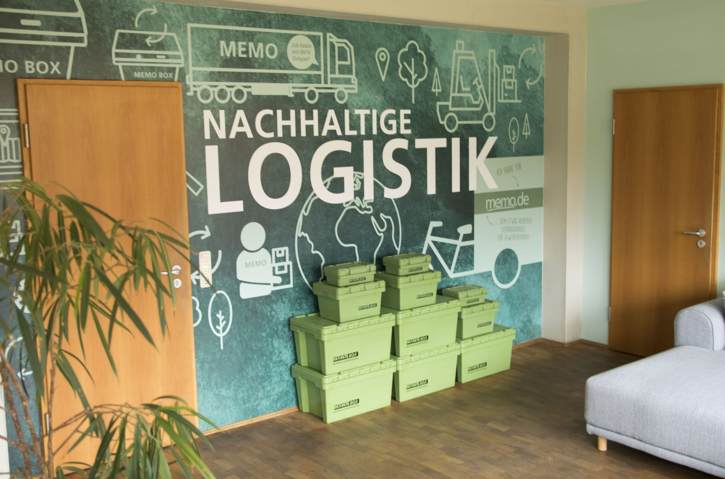 Grüne memoBOXen sind neben einer blaugrünen Wand mit Illustrationen und dem Schriftzug „Nachhaltige Logistik“ gestapelt. Holztüren und ein Sofa sind zu sehen und vermitteln den Eindruck eines umweltfreundlichen Arbeitsbereichs in der memo AG.