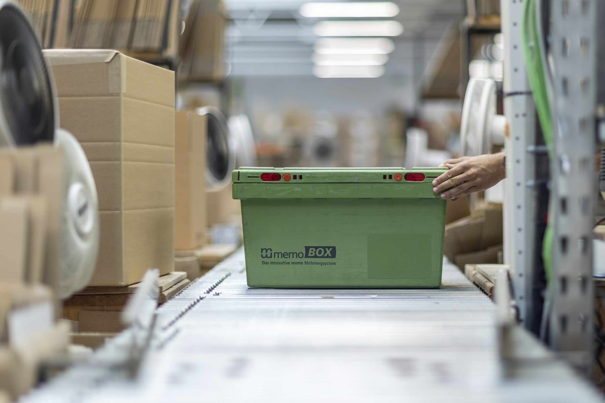 Eine Hand bewegt eine grüne memoBOX auf einem Förderband in einem Lagerhaus. Um die Box herum stehen Stapel von Kartons. Die Szene wirkt geschäftig und industriell.
