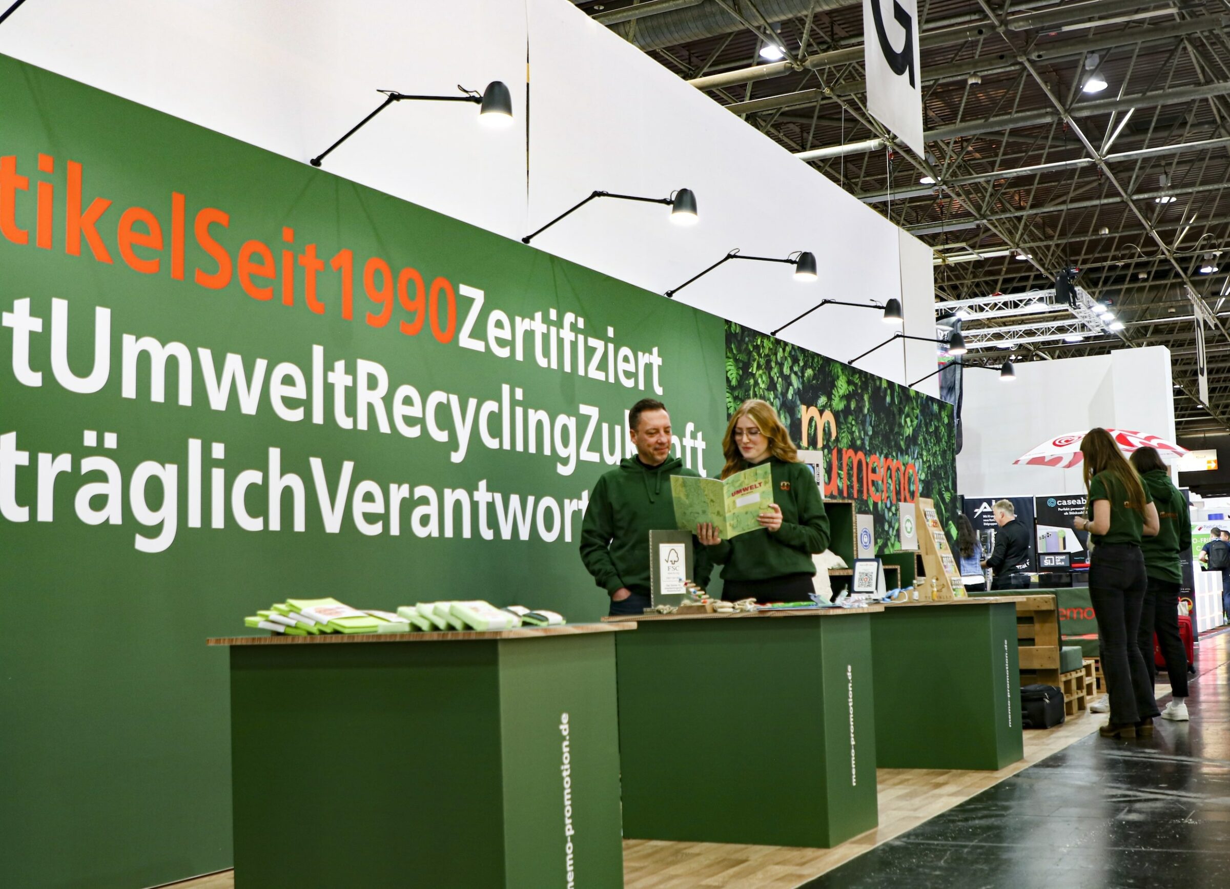 Zwei Personen stehen an einem grünen Messestand der memo AG mit Bannern zum Thema Nachhaltigkeit. Sie begutachten umweltfreundliche Produkte und schaffen so eine informative und bewusste Atmosphäre.