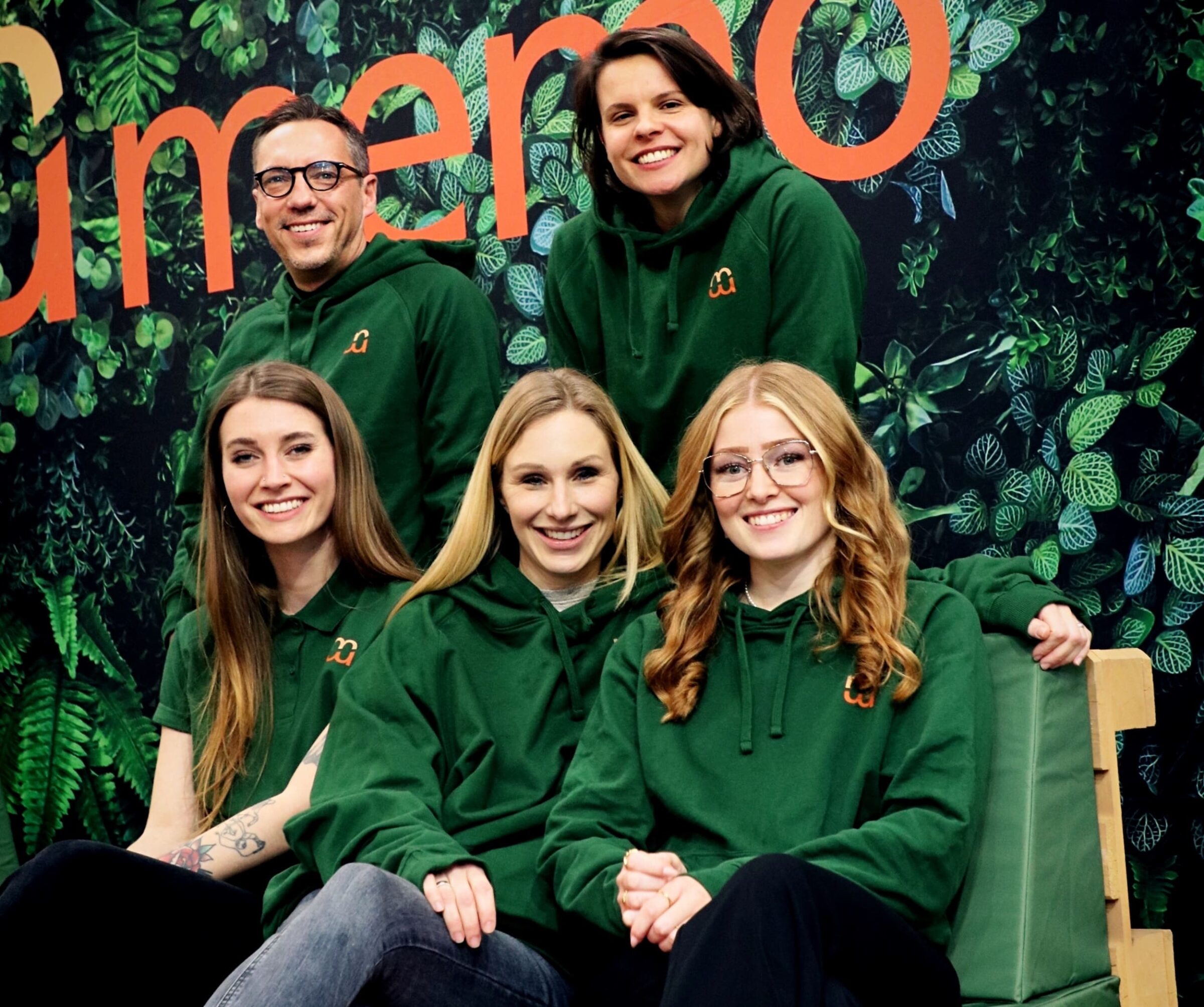 Eine Gruppe von fünf Personen von der memo AG, lächelnd, trägt passende grüne Kapuzenpullis vor einem grünen Hintergrund. Der Ton ist freundlich und harmonisch und vermittelt Teamgeist.