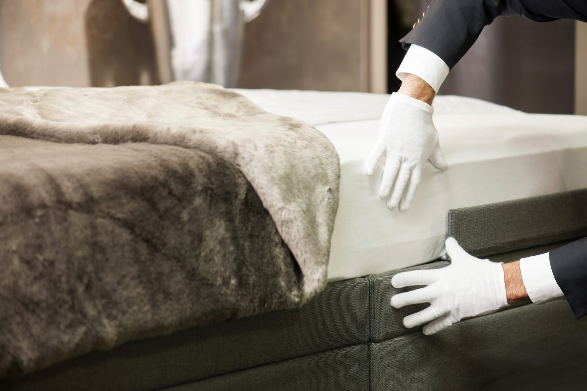 Ein Hotelmitarbeiter mit weißen Handschuhen glättet ein ordentlich gemachtes SWISSFEEL-Bett mit einer hellbraunen Kunstfelldecke. Die Umgebung vermittelt Luxus und Liebe zum Detail.
