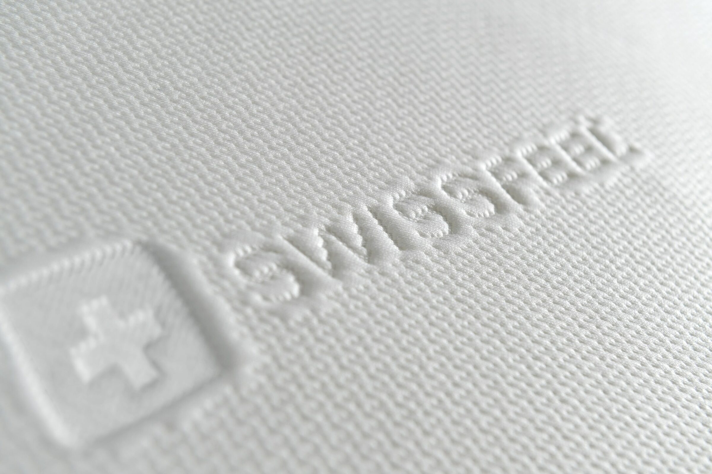 Weißer strukturierter Stoff einer Matratze mit dem Wort „SWISSFEEL” und einem eingeprägten Kreuzsymbol, das ein Gefühl von Weichheit und Premiumqualität vermittelt.