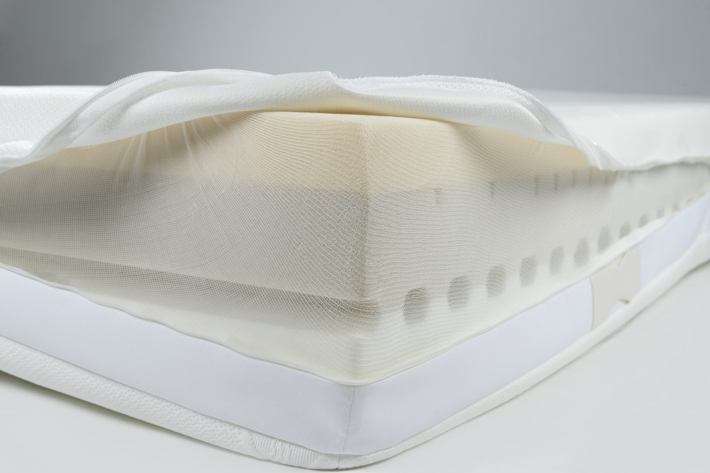 Nahaufnahme einer Ecke einer nachhaltigen SWISSFEEL-Matratze, die die Schichten zeigt. Der obere Stoff ist zurückgeschlagen und gibt den Blick auf eine weiße Schaumstoffschicht mit Perforationen frei. Sauberes und modernes Erscheinungsbild.