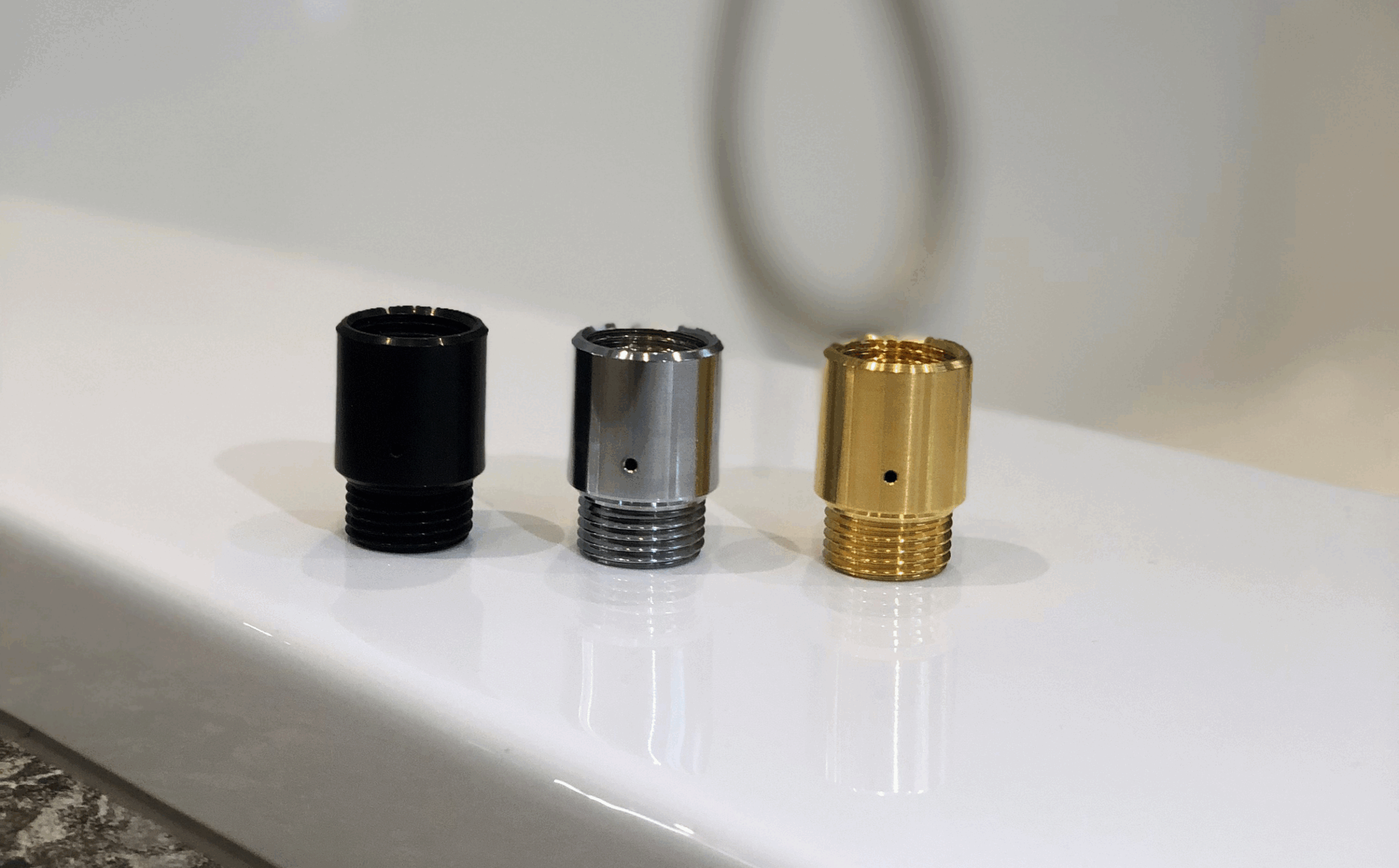 Drei Ecowaterjet Adapter in Schwarz, Silber und Gold sind auf einer weißen Fläche aufgereiht. Der Hintergrund ist leicht unscharf, wodurch eine ruhige, minimalistische Atmosphäre entsteht.