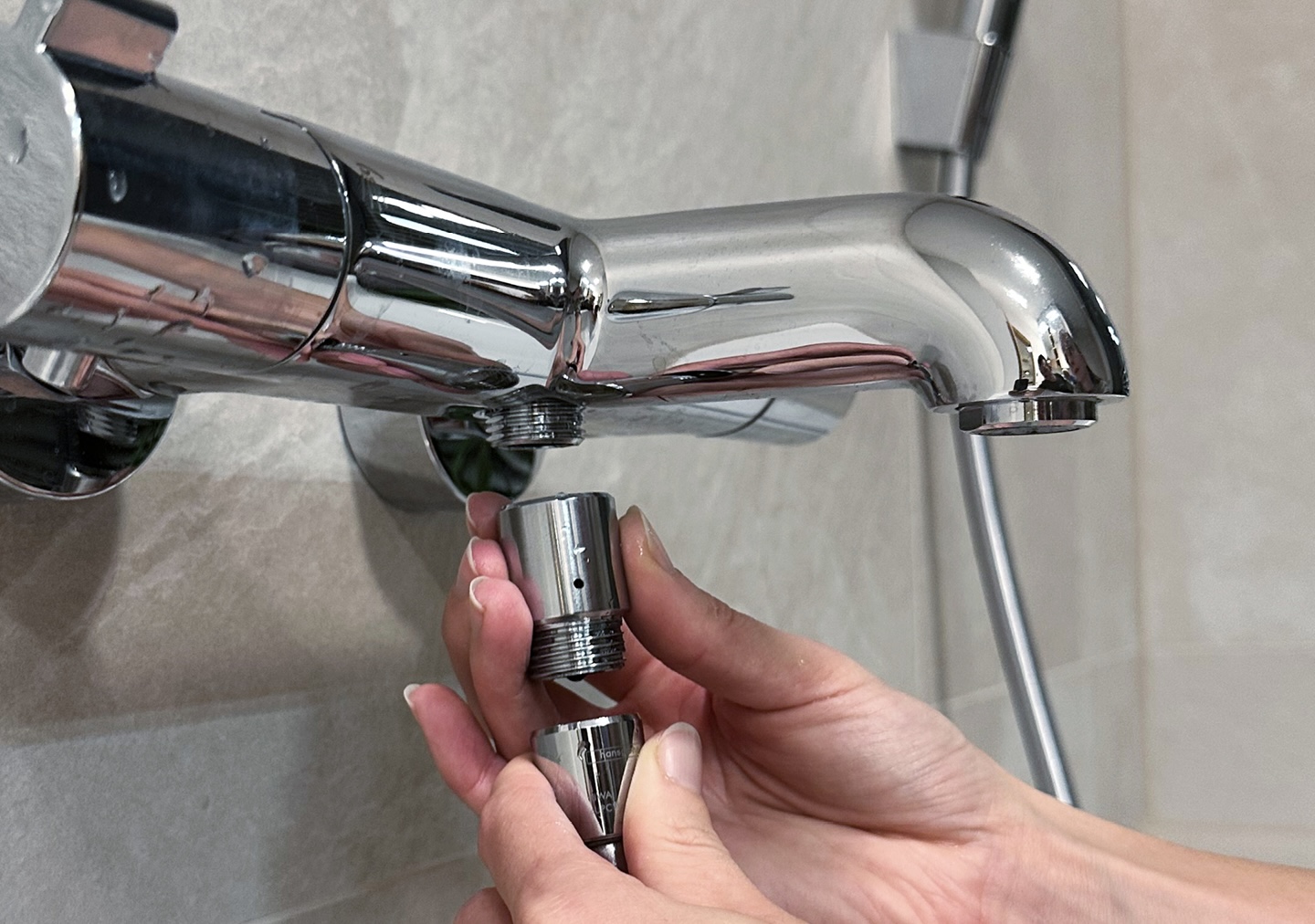 Hände montieren den Ecowaterjet an einem glänzenden Chrom-Waschbeckenhahn vor einer beige gefliesten Wand. Die Szene deutet auf eine Reparatur oder Wartung im Rahmen von Heimwerkerarbeiten hin.