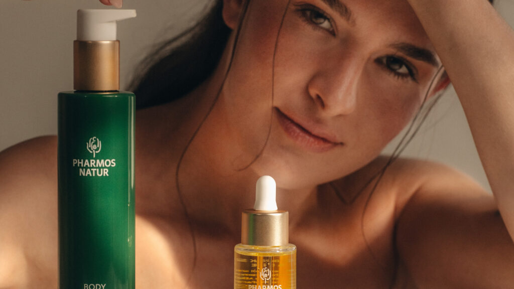 Eine Frau lächelt sanft und hält ihre Hand über ihren Kopf. Vor ihr stehen zwei Hautpflegeprodukte von Pharmos Natur in eleganter grün-goldener Verpackung, die eine ruhige und luxuriöse Atmosphäre schaffen.