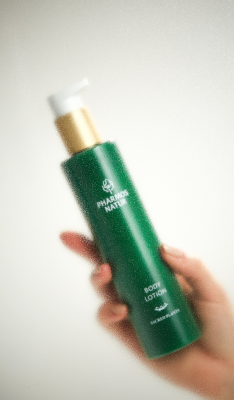 Bild einer grünen Flasche mit der Aufschrift „Pharmax Naturals Body Lotion” von Pharmos Natur mit einer goldenen Pumpe, die von einer Hand gehalten wird. Der Hintergrund ist neutral.