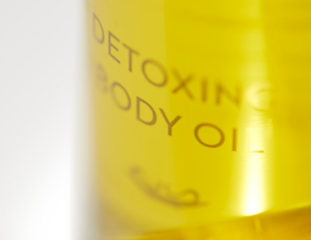 Nahaufnahme einer Flasche mit der Aufschrift „Detoxing Body Oil“. Die Flüssigkeit darin erscheint goldgelb und vermittelt ein frisches, sauberes und beruhigendes Gefühl.