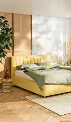 Ein gemütliches Schlafzimmer verfügt über ein gelbes Polsterbett aus der nachhaltigen LA SILVA-Kollektion von Segmueller (Made in Germany) mit grüner Bettwäsche, neben einer grünen Topfpflanze. Heller Holzboden und ein weicher Teppich sorgen für zusätzliche Wärme.