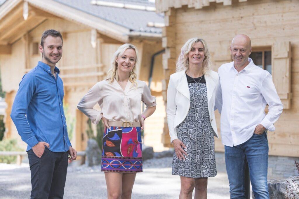 The Moser family stands smiling in front of a chalet outdoors. In the middle are two women in dresses, flanked by men in casual shirts. The atmosphere is cheerful and lighthearted.
