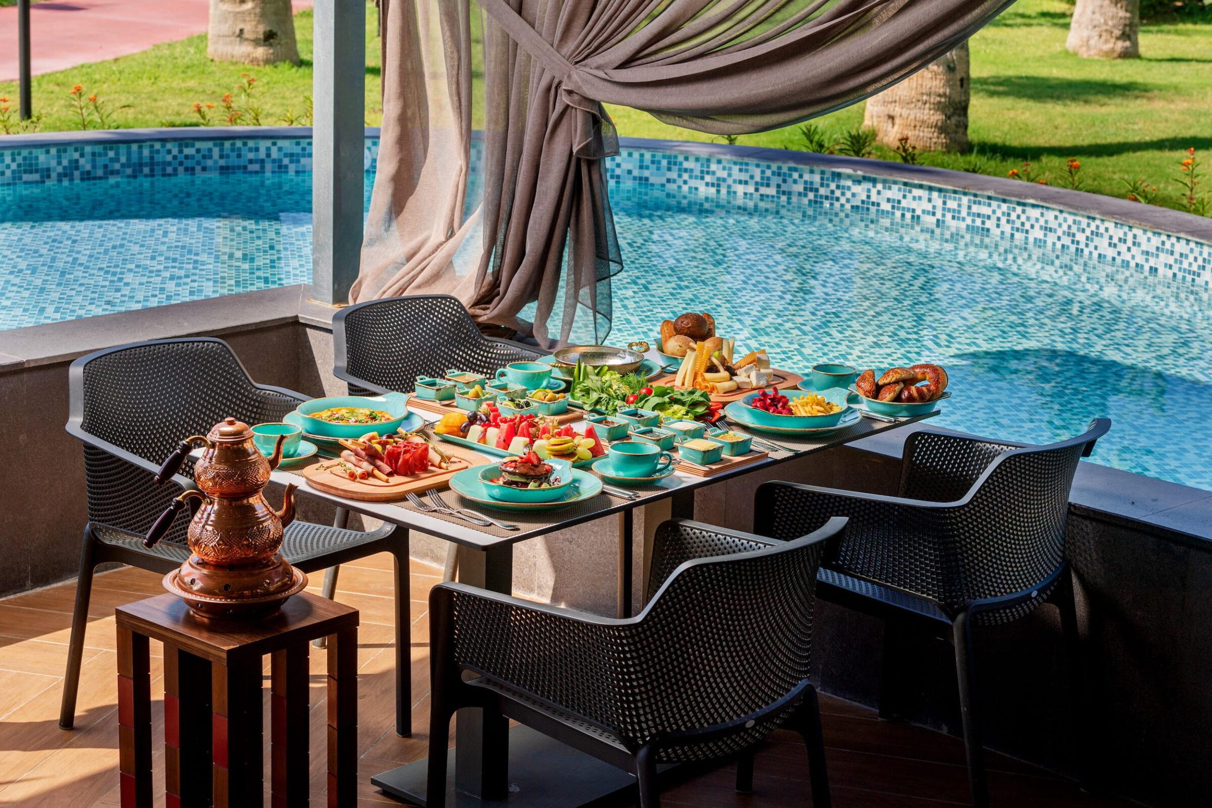 Outdoor dining arrangement by the pool at the Turkish GSTC-certified all-inclusive resort Megasaray Club Belek, with a table full of colorful food and drinks. Black chairs surround the table, and transparent curtains add a touch of elegance.