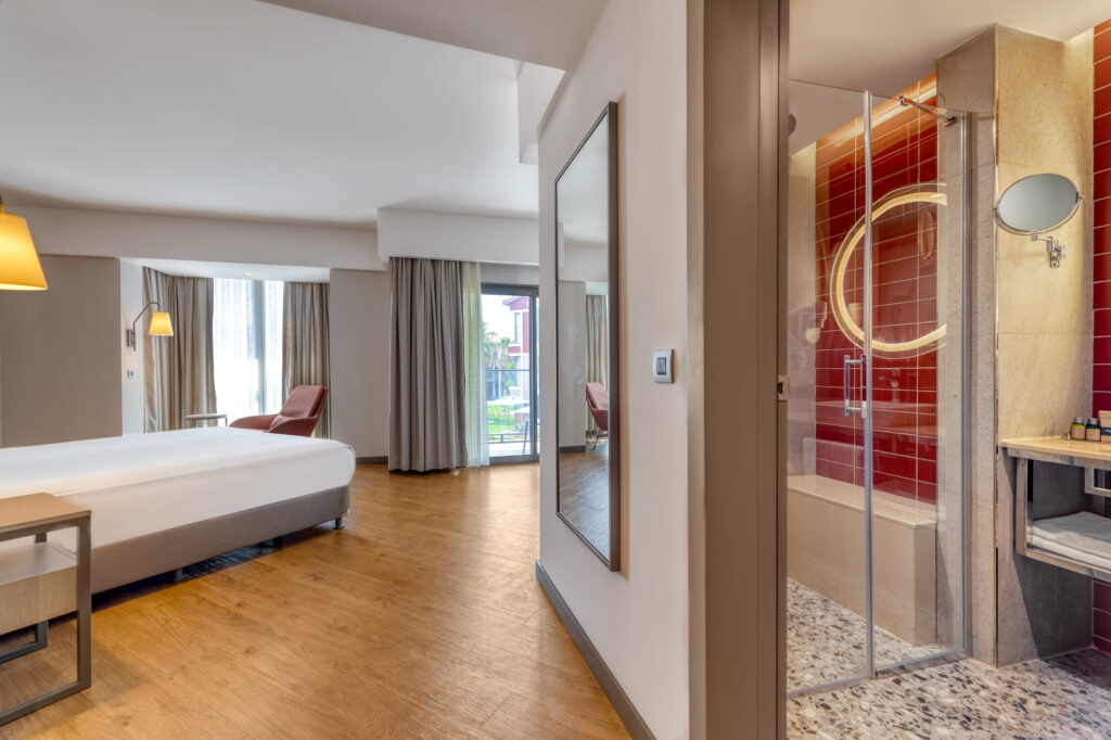 Modern hotel room at the Turkish resort Megasaray Club Belek with wooden flooring, a bed, and a side table. Large window with curtains and a bathroom with red accents and a glass door. Bright and inviting.