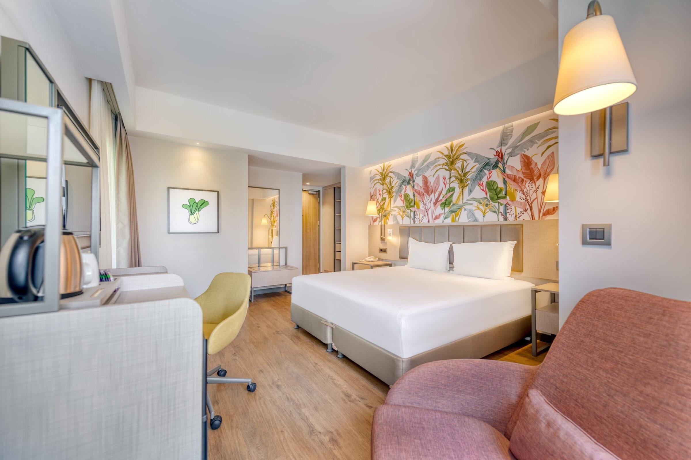 Modern hotel room at the GSTC-certified all-inclusive resort Megasaray Club Belek in Turkey with a cozy atmosphere, large bed, colorful botanical mural, red armchair, and desk with yellow chair.