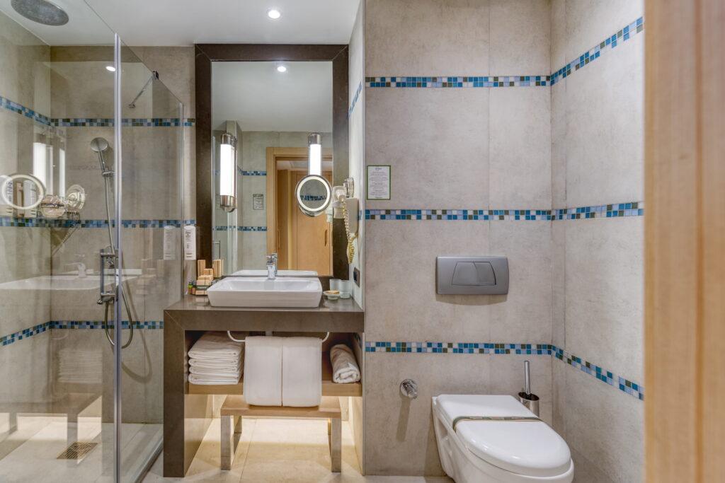 Modern bathroom in a room at the Megasaray Club Belek in Turkey with glass shower, vanity with sink, and wall-mounted toilet. Beige tiles with blue mosaic accents create a clean, elegant look.