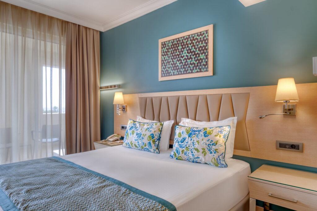 A cozy hotel room at the all-inclusive Megasaray Club Belek resort in Turkey with a double bed, floral pillows, and blue-green bedding. Soft light from wall lamps and sheer curtains creates a peaceful atmosphere.