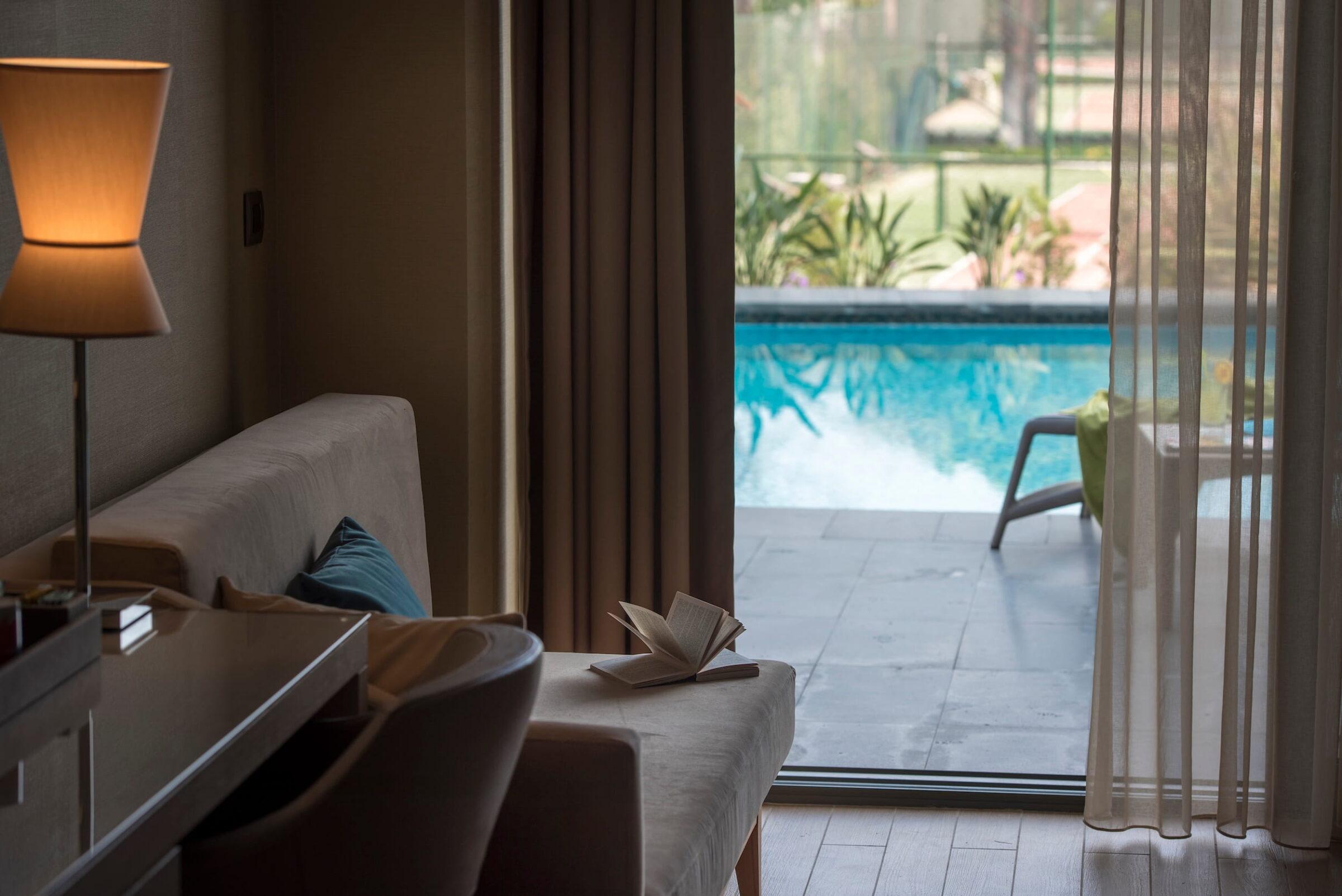 Cozy room at the sustainable Megasaray Club Belek resort in Turkey with a burning lamp, a chair, and an open book on a sofa. Through the transparent curtains, you can see a tranquil pool and garden, creating a peaceful, relaxed atmosphere.