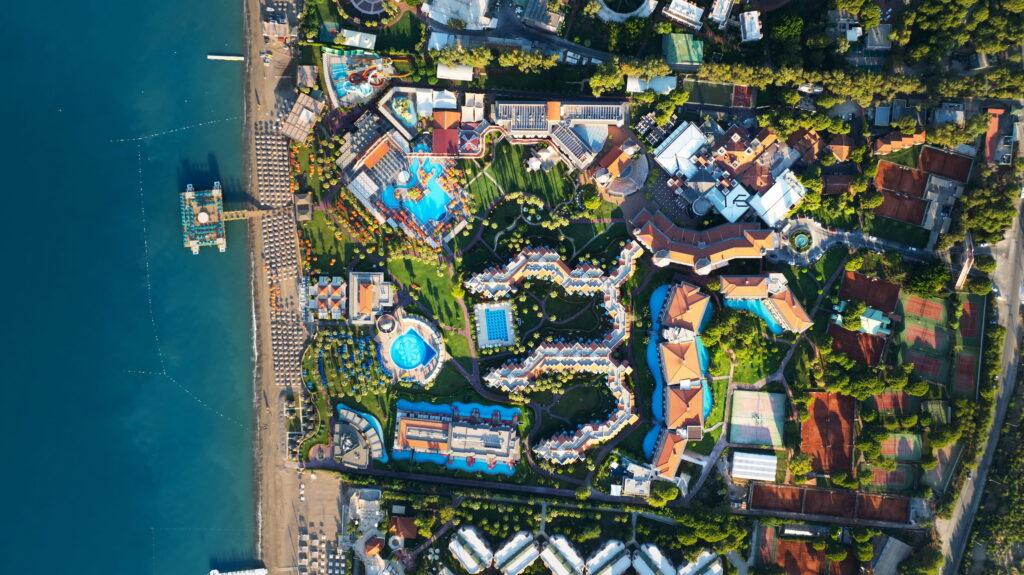 Aerial view of the Megasaray Club Belek beach resort in Turkey with pools, gardens, and colorful buildings. The beach and a pier extend to the left toward the blue sea, creating a lively and relaxed atmosphere.