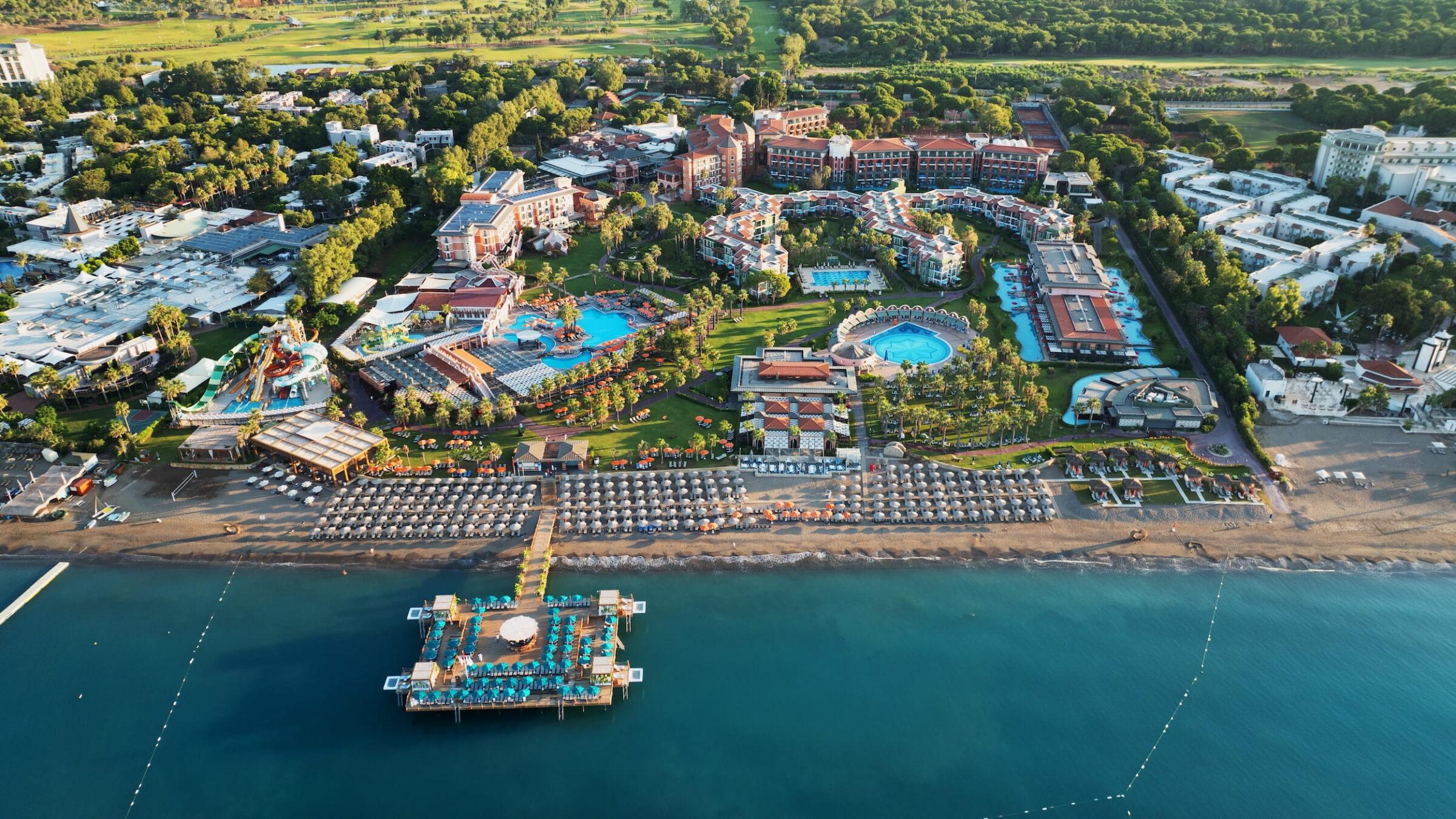 Aerial view of the Megasaray Club Belek coastal resort with several pools, lush gardens, and numerous buildings. A long pier juts out into the calm blue water, while parasols and sun loungers line the sandy beach, creating a relaxed, inviting atmosphere.