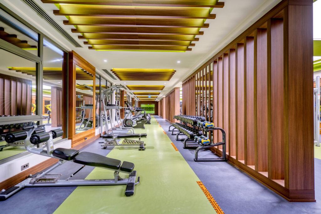 Bright, modern gym at the GSTC-certified all-inclusive resort Megasaray Club belek with mirrored walls, wood accents, and green flooring. Equipped with weights, benches, and exercise machines. Inviting and well-organized space.