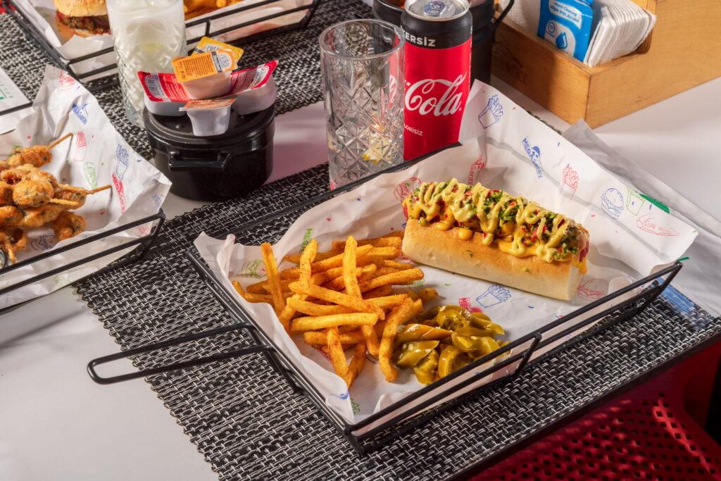 A hot dog with mustard and herbs, served with seasoned French fries and sliced jalapeños in a basket. Next to it is a glass, a beverage can, and condiments. Served at the GSTC-certified Megasaray Club Belek in Antalya, Turkey.