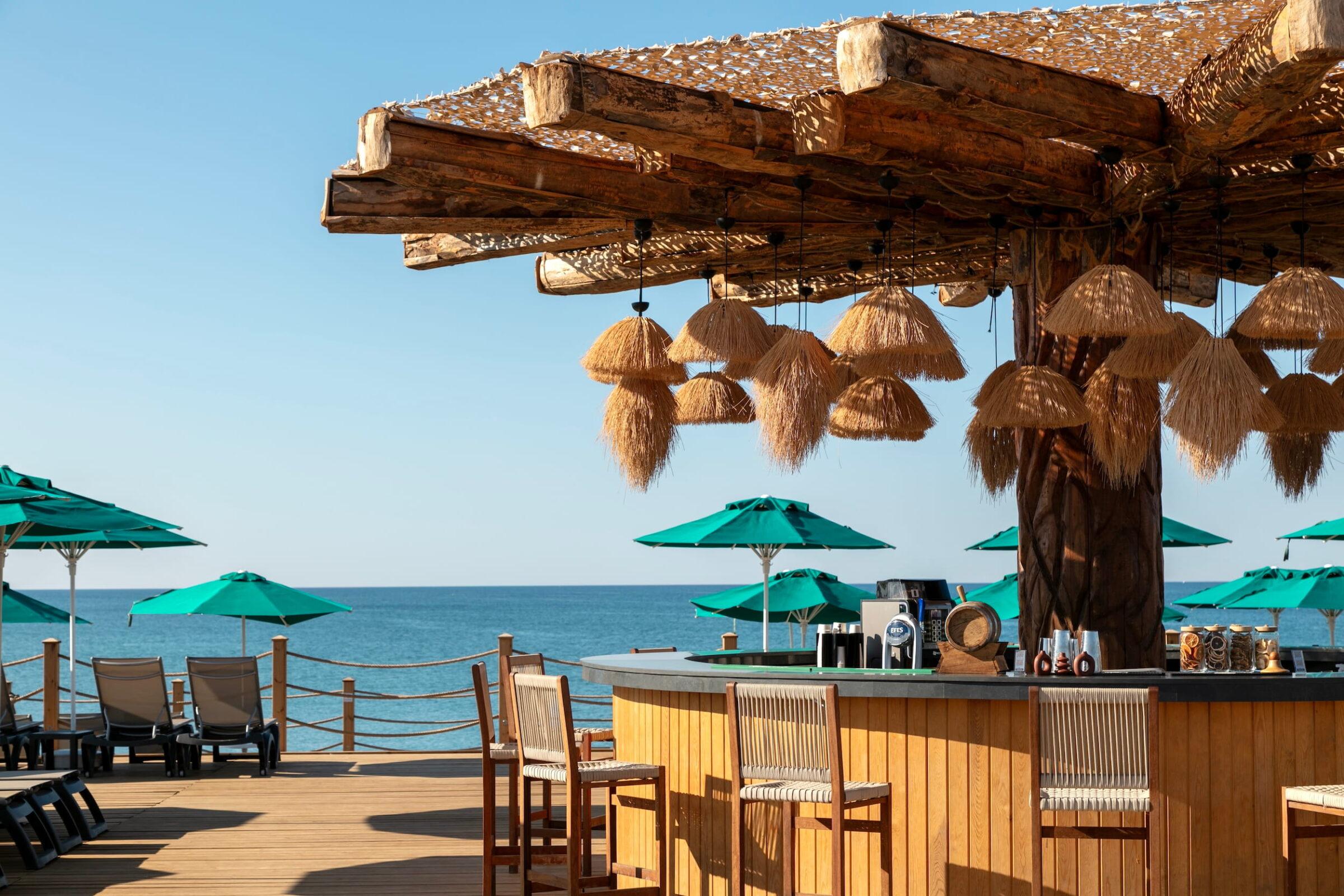 Wooden beach bar at the GSTC-certified Megasaray Club Belek resort in Antalya with straw lamps and wooden stools, overlooking the calm sea. Green parasols and deck chairs create a relaxed, tropical atmosphere.