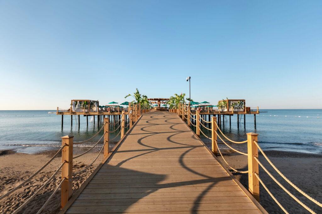 A luxurious wooden walkway at the GSTC-certified Megasaray Club Belek resort stretches out over the calm blue waters and is equipped with rope railings on both sides. The walkway leads to a pavilion with plants and turquoise parasols under a bright blue sky.
