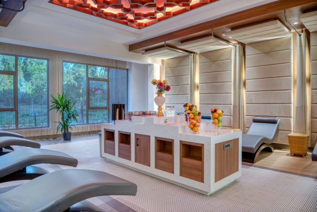 Luxurious spa lounge with sleek chaise lounges, a central table adorned with fruit bowls and flowers, soft lighting, and large windows with a forest view.
