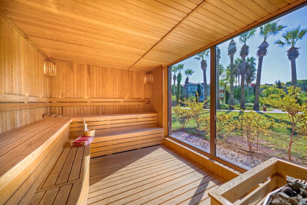 Wood-paneled sauna with benches, large window overlooking the sunny garden of the Megasaray Club Belek with palm trees and shrubs, creating a peaceful and relaxing atmosphere.