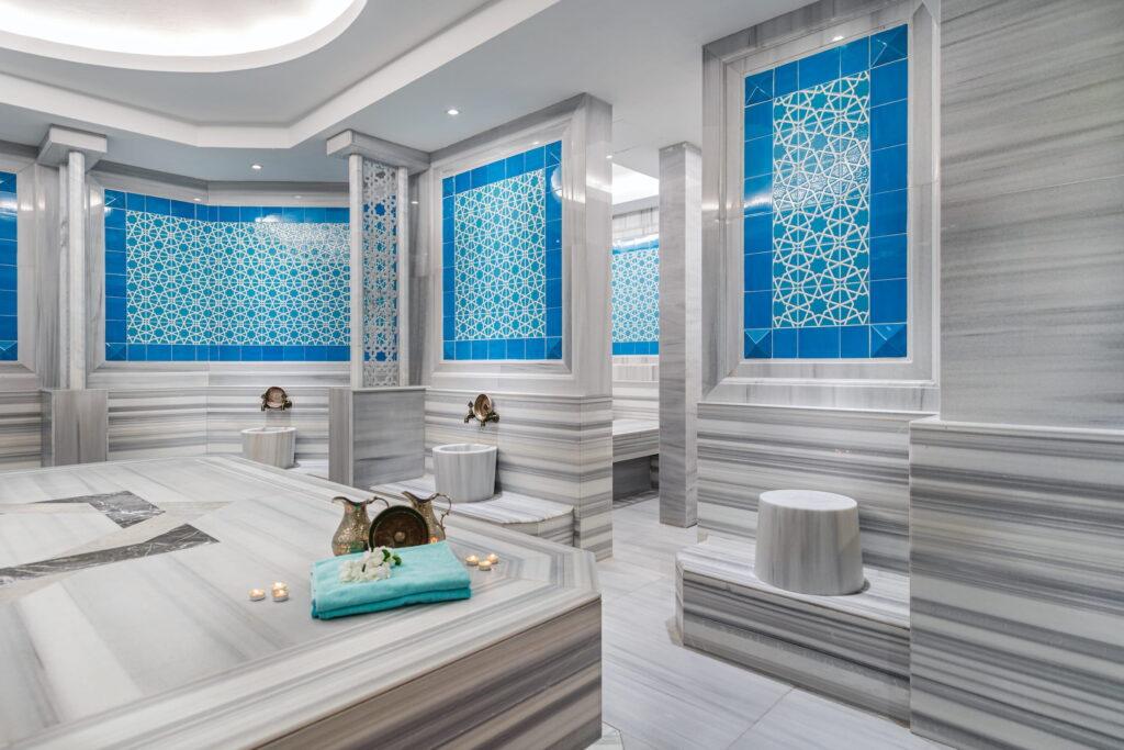 Spa room and Turkish bath at the GSTC-certified Megasaray Club Belek resort, featuring elegant blue and white geometric tiles, marble surfaces, candles, folded towels, brass jugs, and a calm, relaxing atmosphere.
