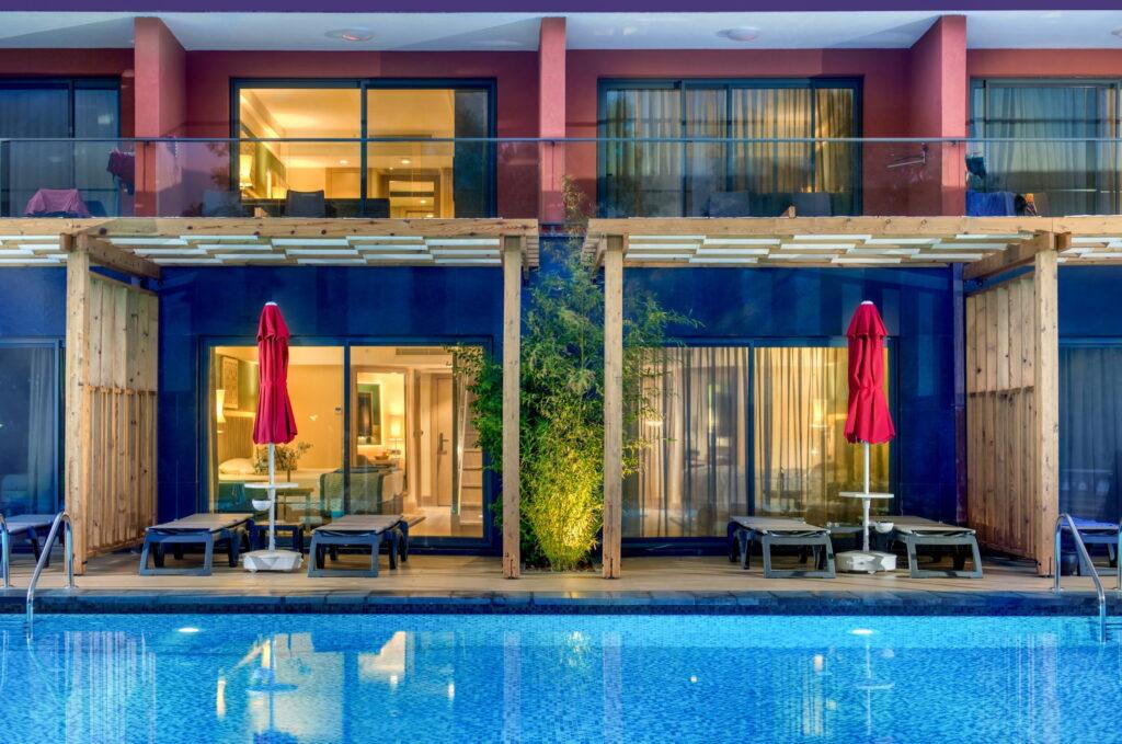 Modern hotel terrace at the GSTC-certified Megasaray Club Belek resort with glass doors leading to the pool. Two red parasols and sun loungers flank a central plant under wooden pergolas, creating a tranquil atmosphere.