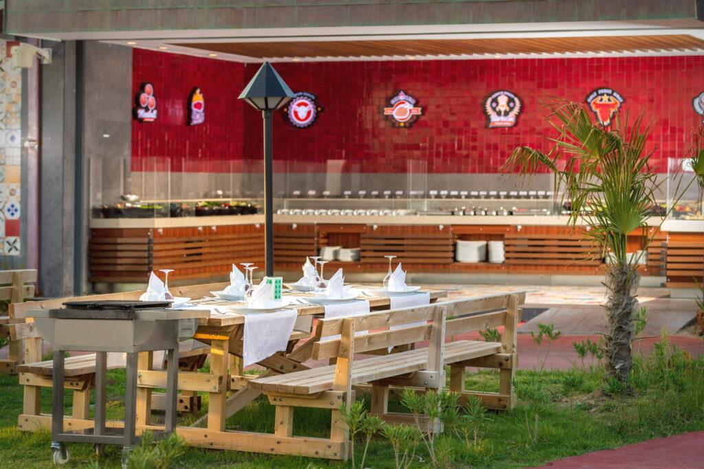 Outdoor area of the GSTC-certified Turkish resort Megasaray Club Belek with wooden tables covered with white napkins and chairs on lawns. A red wall, logos, and a catering station can be seen in the background. Relaxed atmosphere.