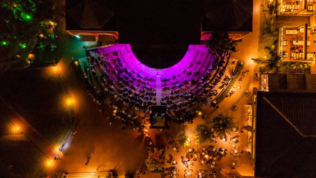 Aerial view of an open-air amphitheater illuminated by purple lights, surrounded by people sitting at tables. Warm, atmospheric lighting creates a festive atmosphere.