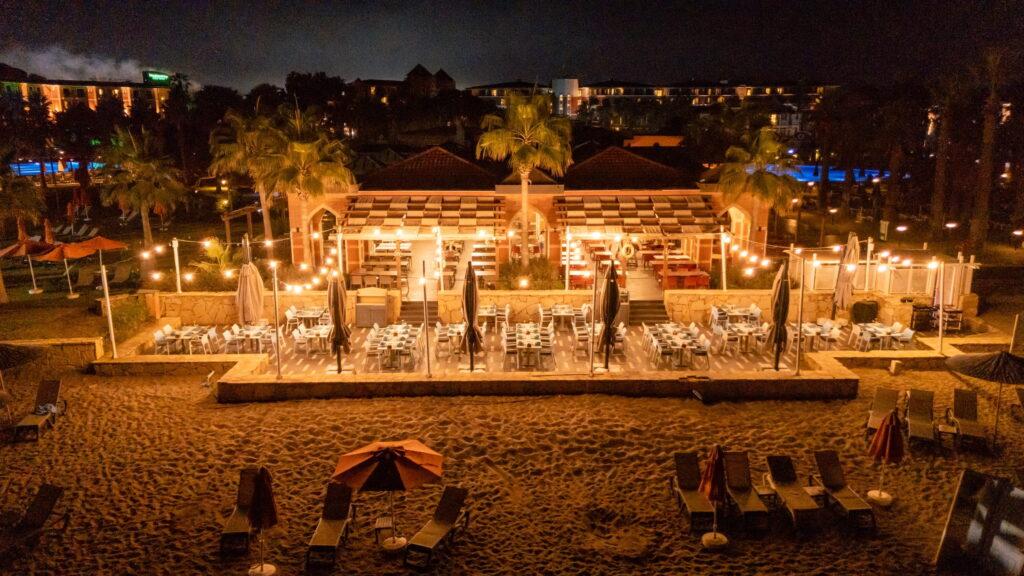 Nighttime beach scene with a warmly lit beach restaurant. Palm trees and fairy lights create a cozy atmosphere. Empty tables and chairs face the sandy beach.