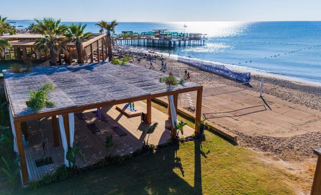 Coastal landscape from the Megasaray Club Belek in Antalya, Turkey, featuring a wooden beach pavilion surrounded by palm trees, overlooking a sand volleyball court and the calm blue sea under a bright blue sky.