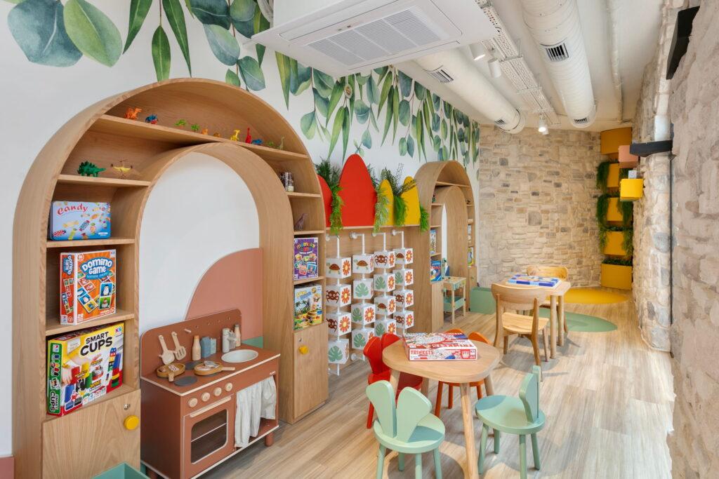 Bright children's playroom in the kids' club at Megasaray Club Belek with leaf wallpaper, wooden shelves, educational games, and a play kitchen. Colorful chairs and tables create a cozy atmosphere.
