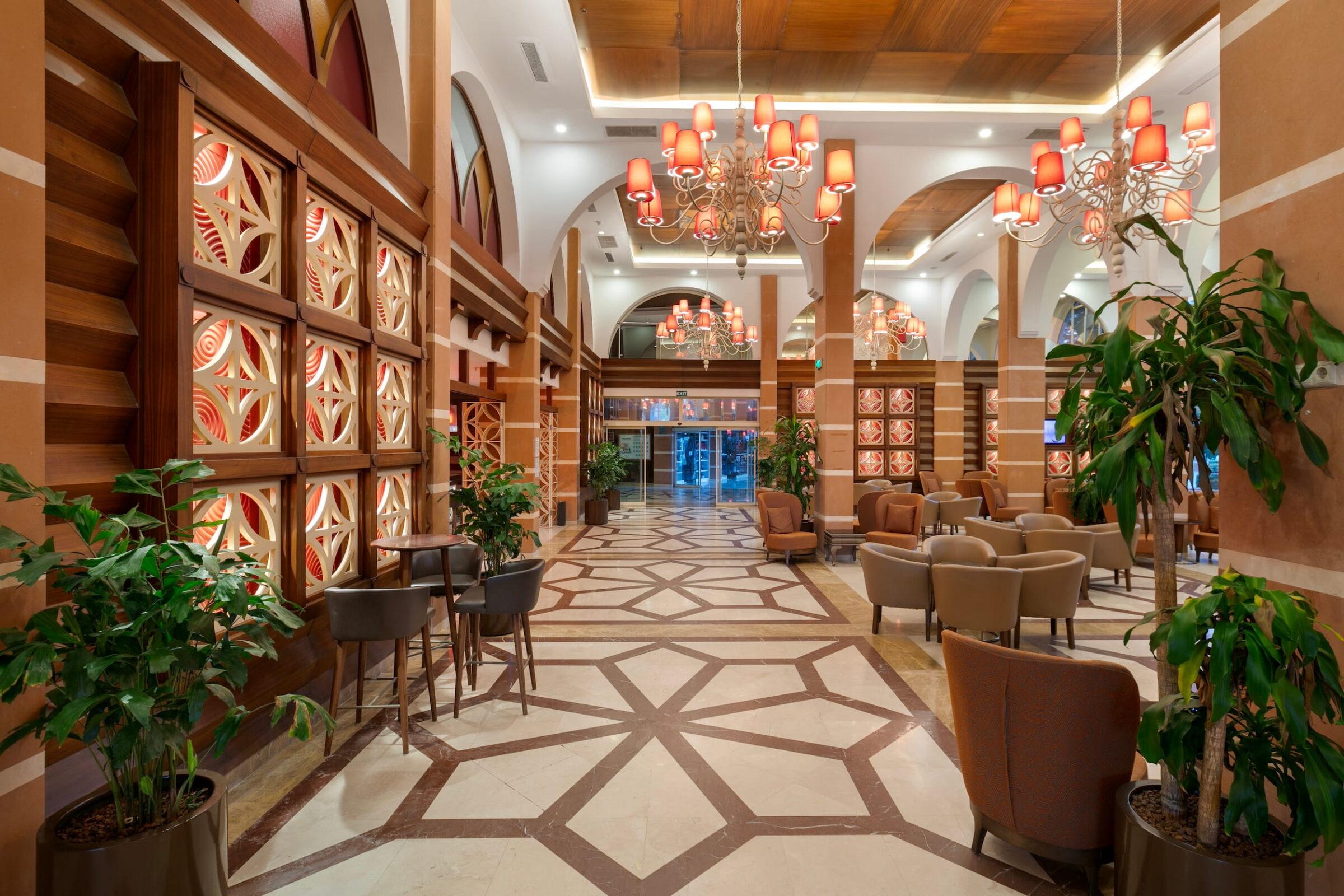 Luxurious hotel lobby of the GSTC-certified Megasaray Club Belek resort with elegant chandeliers, stylish patterned flooring, comfortable seating, and vibrant indoor plants that create a warm and welcoming atmosphere.