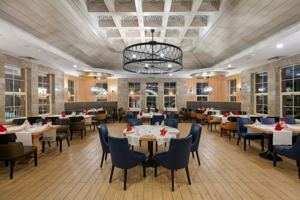 Spacious, elegant restaurant interior in the main restaurant of the all-inclusive Megasaray Club Belek resort, featuring wooden floors and high ceilings. Blue chairs surround tables set with white tablecloths and red napkins, creating a welcoming atmosphere.