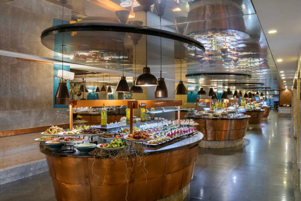 Luxurious buffet at the Turkish all-inclusive resort Megasaray Club Belek with various regional dishes served on round wooden tables. Pendant lights create a warm atmosphere in the elegant, mirrored dining room.