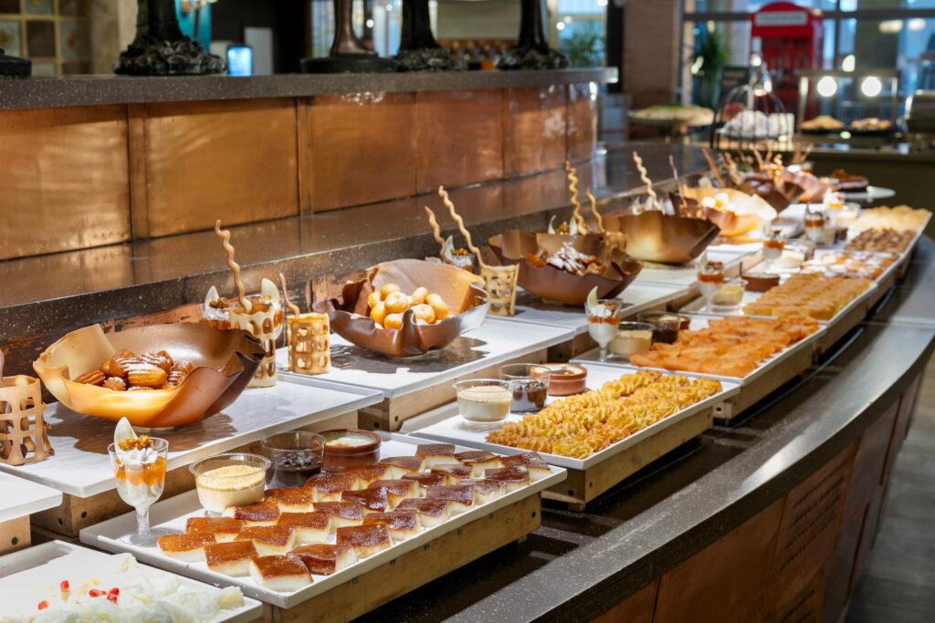 A lavish dessert buffet in the main restaurant of the sustainable Megasaray Club Belek resort in Turkey offers a selection of sweet treats such as cakes, pastries, and puddings, arranged in elegant bowls and on trays to create a warm, inviting atmosphere.