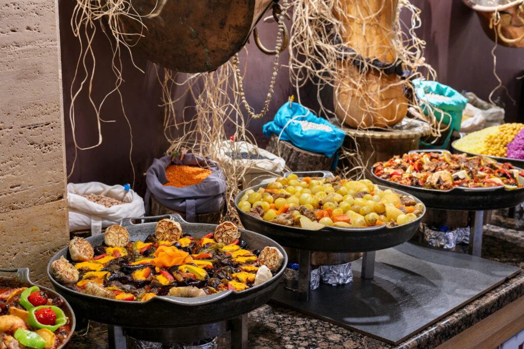 A colorful buffet in the main restaurant of the sustainable Megasaray Club Belek resort in Antalya, Turkey, featuring colorful dishes made with vegetables, beans, and meat in large pans. The rustic décor with hanging grapevines creates a cozy, inviting atmosphere.