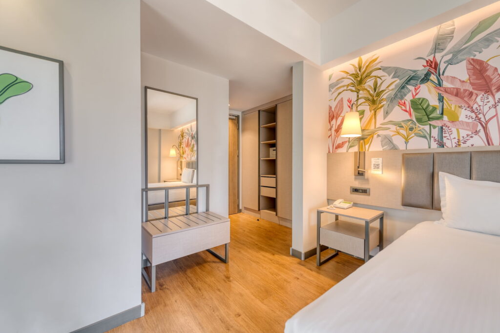 Bright hotel room at the sustainable Megasaray Club Belek in Turkey with wooden floors, a bed, a bedside table with lamp, tropical-style wall decorations, a mirror, and an open wardrobe. Cozy and inviting.