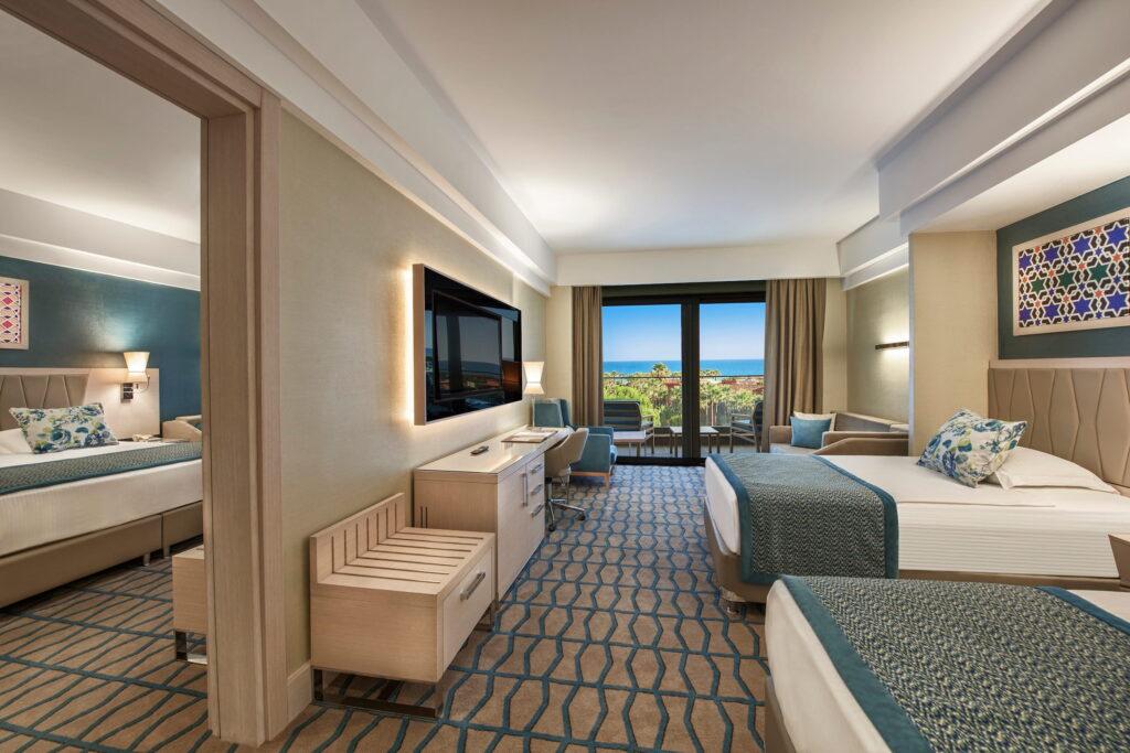 Modern hotel room at the GSTC-certified Megasary Club Belek resort in Antalya with two single beds and sea views. Neutral colors, geometric carpet, wall decorations, large TV, and a balcony with chairs provide additional tranquility.