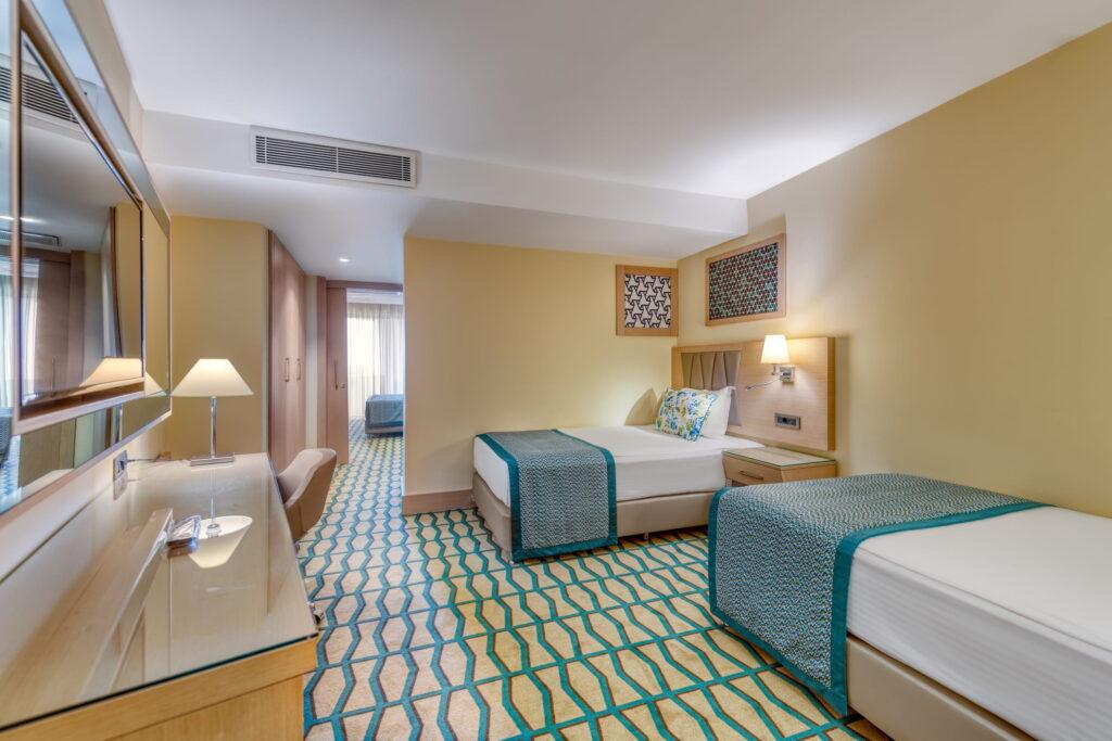 A hotel room at the all-inclusive Megasaray Club Belek resort in Turkey with two single beds, blue-green accents on the bedding and carpet, a wooden desk with a lamp and soft lighting that creates a soothing atmosphere.