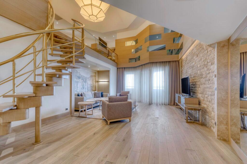 Spacious, modern living room in the Pasha Suite at Megasaray Club Belek in Turkey with spiral staircase, wooden floors, and an accent wall made of stone. Comfortable seating, large TV, and soft daylight through high curtains.