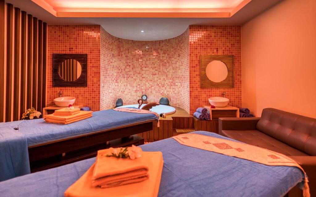 Luxurious spa room at Megasaray Club Belek with two massage tables covered with blue towels, decorated pink mosaic wall, round mirrors, and warm ambient lighting. Relaxing atmosphere.